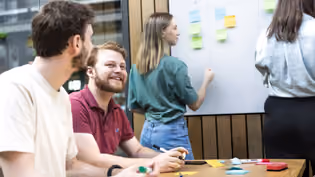 Computer Science students collaborate on a project, with one writing on a sticky-note-covered whiteboard and others discussing.
