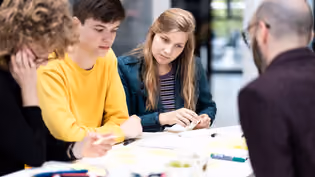 A group of students or professionals engaged in a collaborative discussion or workshop. They appear to be brainstorming or working on a project, with sticky notes and other materials on the table.