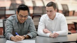 Two Business IT students sit at a table in a classroom, one writing on paper while the other observes and engages in discussion.