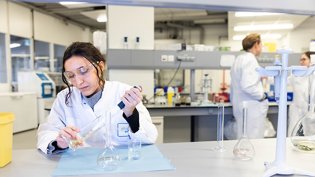 Student in a laboratory setting, wearing a lab coat and safety goggles, working with glassware and a pipette. The focus is on conducting experiments, likely related to water technology or chemical analysis, with other lab personnel visible in the background.