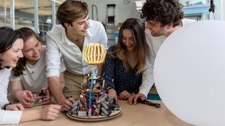 Five students work together on an innovative technical project in a makerspace. They smile and look enthusiastically at a home-made robot with an illuminated, spherical structure and complex wiring. On the table are tools and electronic components. To the right of the robot is a large white balloon. The environment has a creative and technical feel with workbenches and equipment in the background.