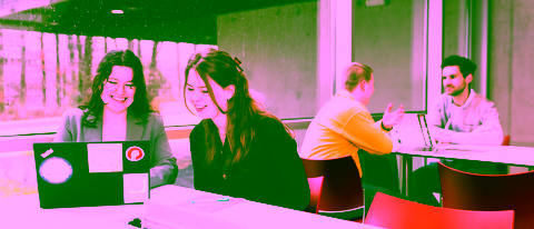 Two female students public administration smile while working on a laptop, with two men talking in the background by a large window on a rainy day.