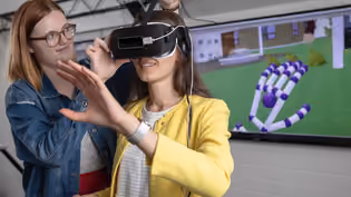 A student wearing a yellow jacket uses a virtual reality headset while another student adjusts the device. A screen in the background displays a 3D simulation of a hand, showcasing interactive VR technology.