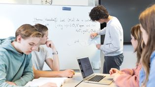 An Applied Mathematics student writes sums on a board, while four other students sit at a table watching.