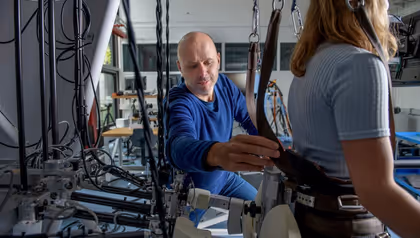 Researcher adjusting straps of a wearable exoskeleton system on a participant in a laboratory filled with cables and mechanical equipment.