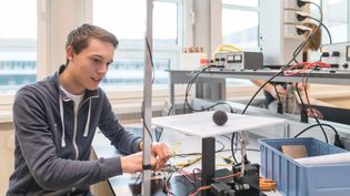 Drie studenten werken samen aan een natuurkundig experiment in een klaslokaal. Een laptop op tafel toont een presentatie met de titel "Method of Images" en diagrammen van elektrische veldlijnen. De studenten bestuderen een doorzichtige behuizing met elektronische componenten en een luidspreker. Eén student leunt op de tafel, terwijl de andere twee actief betrokken zijn bij het experiment.