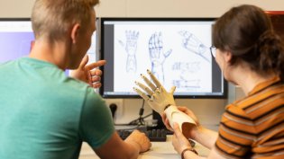 Two people in a design studio discuss a 3D printed prosthetic hand. In the background, computer screens show technical drawings and designs of the prosthesis