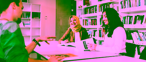 A diverse group of three students around a table in a library, laughing and talking. The table is filled with open books and in the background are shelves full of books