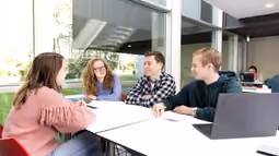 Four mathematics students are sitting together at a table in a modern study space, engaged in discussion.