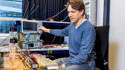 A teacher operates electronic testing equipment in a lab, surrounded by connected devices and wires on the table.