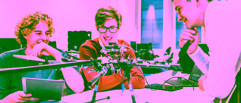 Three students collaborating on a robotics project, examining a large drone with exposed wiring and components in a modern laboratory setting. One holds a tablet, while the others observe and discuss the design