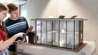 Two Electrical Engineering students examine a detailed architectural model of a modern building, with one using a tablet.
