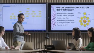 Four Business IT students discuss a project in a meeting room with screens showing a flowchart and a slide on Enterprise Architecture.