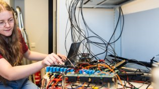 A student works on a complex setup of electronic components and wires arranged on a desk in a technical lab environment.