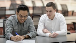Two Business IT students sit at a table in a classroom, one writing on paper while the other observes and engages in discussion.