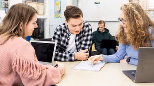 Three mathematics students are working together on an assignment, while a fourth student is sitting in the background with a laptop.