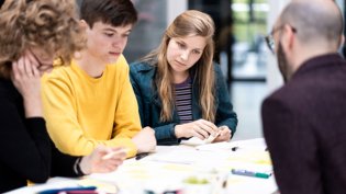 A group of students or professionals engaged in a collaborative discussion or workshop. They appear to be brainstorming or working on a project, with sticky notes and other materials on the table.