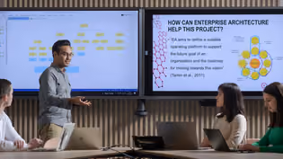 Four Business IT students discuss a project in a meeting room with screens showing a flowchart and a slide on Enterprise Architecture.
