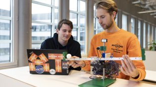 Two young men working on an engineering project in a modern, well-lit workspace. One, wearing an orange sweatshirt, is adjusting a small robotic system with electronic components and a mechanical structure resembling a balance or control system. The other, in a dark hoodie, is observing while working on a laptop covered in stickers.