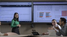 Four Business IT students collaborate in a meeting room with two large screens showing code and data visualisations.