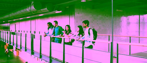 A group of students standing on an indoor walkway, leaning against the railing and looking out over the open space below.