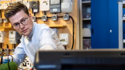 An electrical engineering student works in a lab with various components and equipment, focusing on a hands-on technical task.