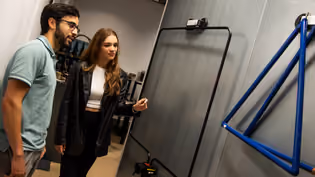 Two Communication Networks students examine a wall-mounted antenna, with one pointing at it during a discussion.