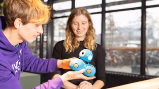 Interaction Technology students engage with a small blue robot in a bright room with large windows, emphasizing hands-on learning.