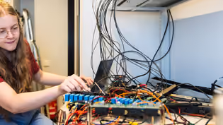 A student works on a complex setup of electronic components and wires arranged on a desk in a technical lab environment.