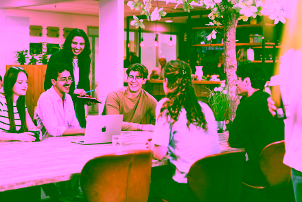 A group of master's students gathered around a table, laughing while looking at a laptop screen.