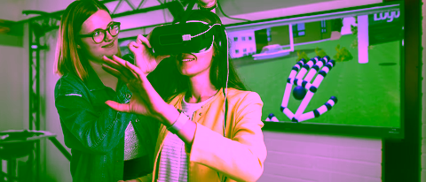 A student uses a virtual reality headset while another student adjusts the device. A screen in the background displays a 3D simulation of a hand, showcasing interactive VR technology