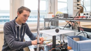 Drie studenten werken samen aan een natuurkundig experiment in een klaslokaal. Een laptop op tafel toont een presentatie met de titel "Method of Images" en diagrammen van elektrische veldlijnen. De studenten bestuderen een doorzichtige behuizing met elektronische componenten en een luidspreker. Eén student leunt op de tafel, terwijl de andere twee actief betrokken zijn bij het experiment.