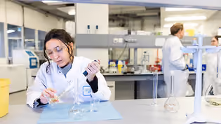 Student in a laboratory setting, wearing a lab coat and safety goggles, working with glassware and a pipette. The focus is on conducting experiments, likely related to water technology or chemical analysis, with other lab personnel visible in the background.