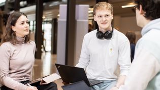 Three students are sitting in an open study space having a conversation. One student has a laptop on their lap and headphones around their neck. Another student is holding a notebook and a pen.