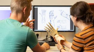 Two people in a design studio discuss a 3D printed prosthetic hand. In the background, computer screens show technical drawings and designs of the prosthesis