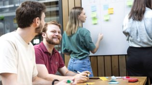 Computer Science students collaborate on a project, with one writing on a sticky-note-covered whiteboard and others discussing.