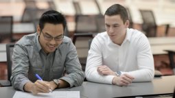 Two Business IT students sit at a table in a classroom, one writing on paper while the other observes and engages in discussion.