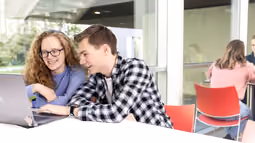 Two math students work on a laptop in a bright, modern space with large windows and others visible in the background.