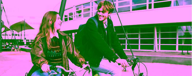 Two students ride bicycles on campus, smiling at each other. The background features a modern university building with large windows and outdoor seating.