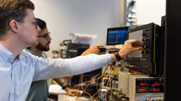 Two students work with electronic equipment and an oscilloscope, monitoring data on screens in a technical lab.
