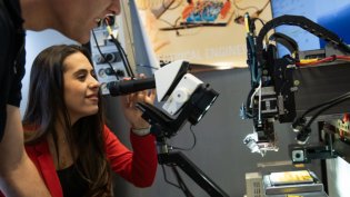 Electrical Engineering student uses a microscope while another observes, surrounded by lab equipment and a computer monitor.