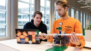 Two young men working on an engineering project in a modern, well-lit workspace. One, wearing an orange sweatshirt, is adjusting a small robotic system with electronic components and a mechanical structure resembling a balance or control system. The other, in a dark hoodie, is observing while working on a laptop covered in stickers.