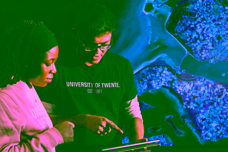 Two students standing in front of a large digital map display, reviewing data together on a tablet. The screen behind them shows colourful satellite imagery, and both students wear University of Twente clothing, suggesting a collaborative geospatial or remote‑sensing learning activity.
