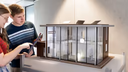 Two Electrical Engineering students examine a detailed architectural model of a modern building, with one using a tablet.