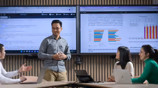 A student gives a presentation on data analysis, while three others engage with the content shown on two large screens.