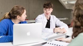 Three students are sitting at a table with laptops and books