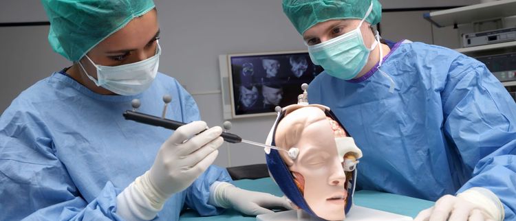 Two people in lab coats during surgical training with an anatomical head model in a clinical setting.