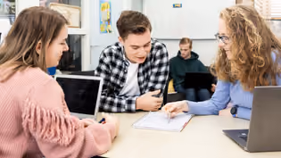 Three math students work at a table with laptops and notebooks, while two others are seen in the background.