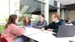 Four mathematics students are sitting together at a table in a modern study space, engaged in discussion.