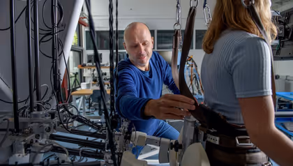 Researcher adjusting straps of a wearable exoskeleton system on a participant in a laboratory filled with cables and mechanical equipment.